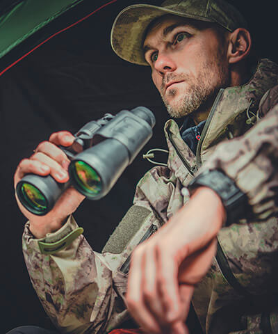 Ventajas Seguro del Cazador - Cazador con prismaticos en las manos y ropa de camuflaje junto a su tienda de campaña en el monte Ventajas Seguro del Cazador - Cazador con prismaticos en las manos y ropa de camuflaje junto a su tienda de campaña en el monte